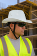 Person wearing a white cowboy hat, sunglasses, and a high-visibility vest in front of a construction site.